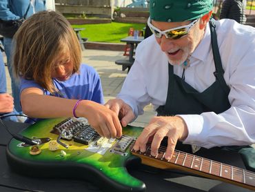 An elderly man teaches a child to play an electric guitar outdoors.