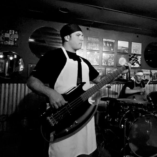 Musicians wearing aprons perform with guitar and drums in a cozy venue.