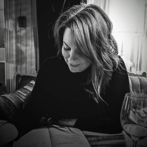 A woman smiles while reading a book at a table with a glass of wine.