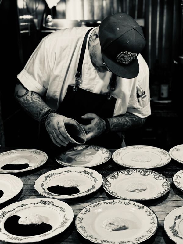Chef with tattoos plating dishes in a dimly lit kitchen.