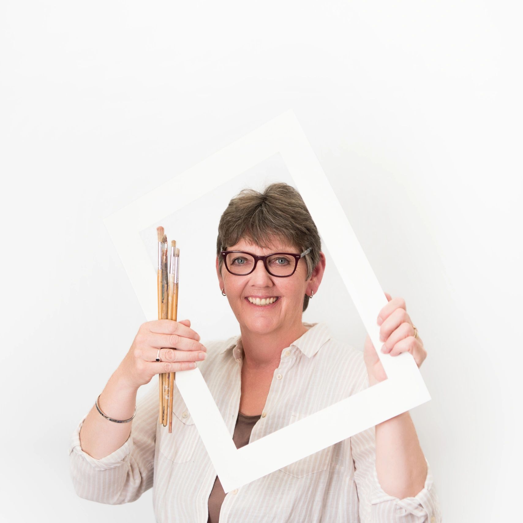 Smiling woman holding paintbrushes and a white picture frame.