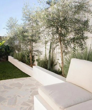 Cozy outdoor seating area with white cushions and lush greenery against a white fence.