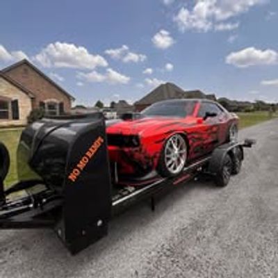 Red muscle car on a flatbed trailer with cloudy sky background.