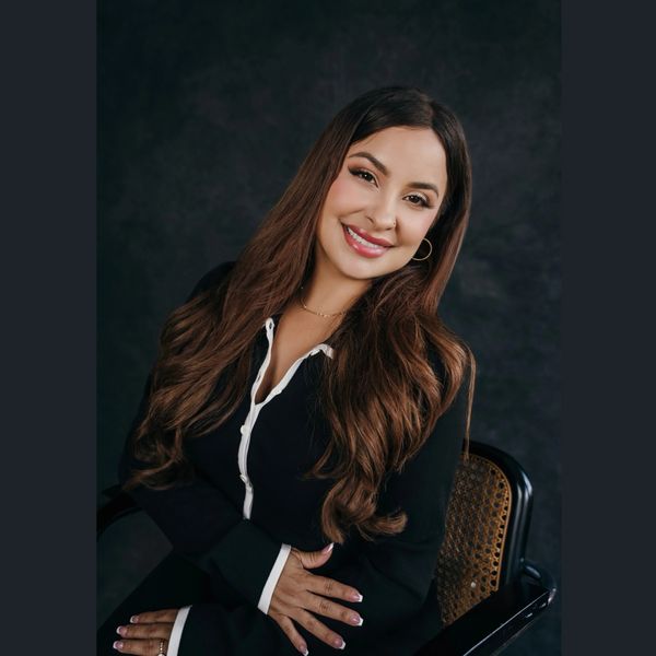 Smiling woman with long brown hair in black outfit sitting on a chair.