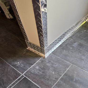 Corner of a wall protected by shiny metal diamond plate trim with dark tiled flooring.