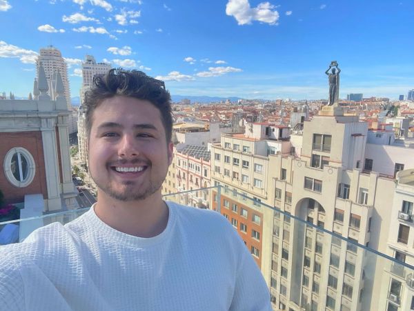 A young man smiling on a rooftop with a cityscape background under a blue sky.