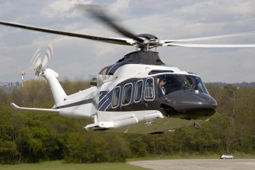 White and black helicopter flying low over a grassy area with a visible pilot inside.