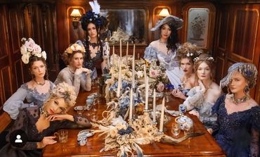 Women in vintage dresses and floral hats around an ornate dining table.