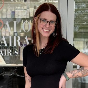 Smiling woman with glasses and tattoos stands outside a hair salon.