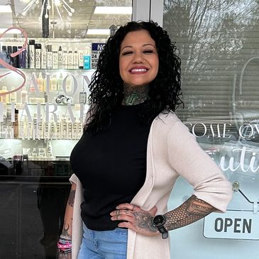 Smiling woman with curly hair and tattoos posing confidently outside a salon.