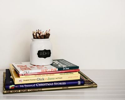 A stack of books against a white wall with a jar full of pencils. A label reads "Read" in chalk.