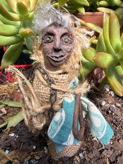 Handcrafted rustic doll wrapped in burlap cloth among green plants.
