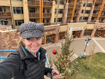Woman taking a selfie near a decorated Christmas tree in an outdoor plaza.