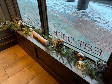 Festive window display with pinecones, lights, and greenery inside a cozy wooden interior.