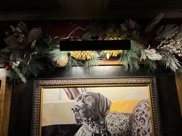 A framed painting of a dog illuminated by a decorative light and greenery above.