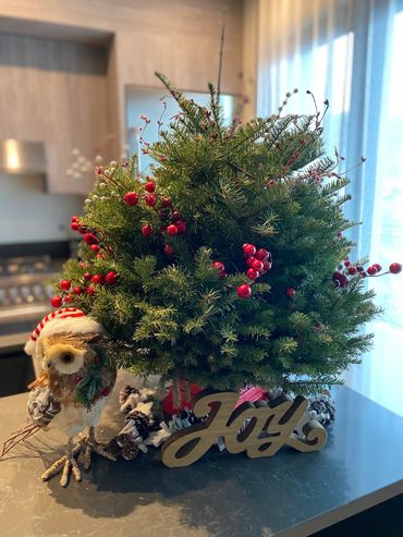 Festive holiday centerpiece with greenery, red berries, an owl decoration, and a wooden 'Joy' sign.
