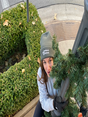 Woman in winter clothes decorating a lamppost with garland outdoors.