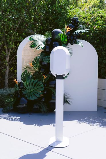White photo booth stand with black balloons and green tropical leaves backdrop outdoors.