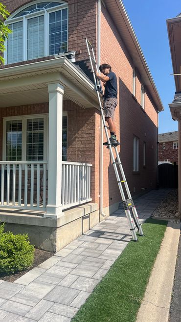 Man on a ladder fixing or inspecting the roof edge of a brick house on a sunny day.