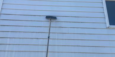 A cleaning brush on a long pole against a dirty white siding wall.