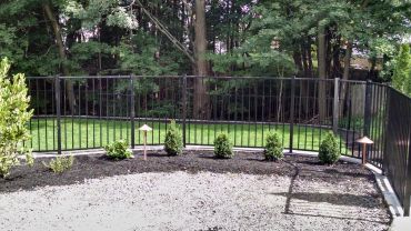 Black metal fence enclosing a green backyard with plants and gravel foreground.