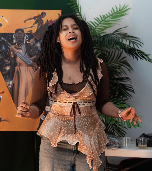 A singer mid-note at a CHECK 1-2 show in Oakland, painted mural and plants behind her.