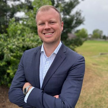 Smiling man in a blue suit standing outdoors with arms crossed.