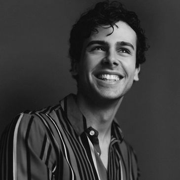 A black & white photo of Ainsley Melham. He is wearing a striped unbuttoned shirt while smiling