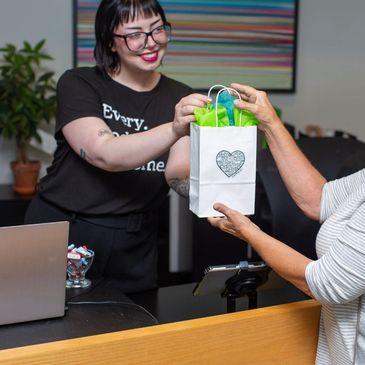 A woman handing a white, green, and blue gift bag over the front desk at East Village Spa. 