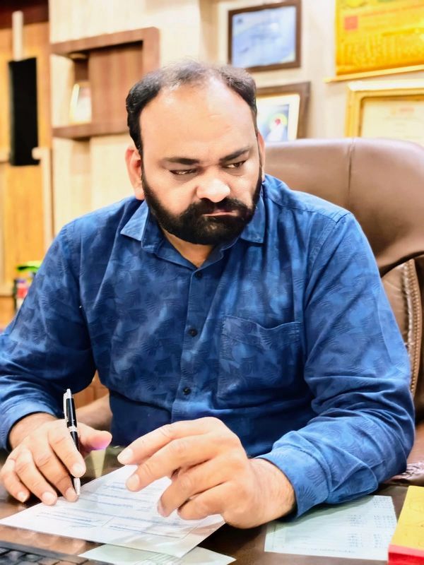 Man in blue shirt sitting at desk, writing on a document with a pen.