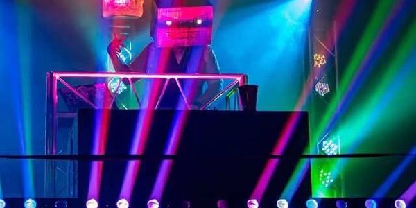 Two DJs with cube-shaped helmets performing on a brightly lit stage with colorful lights.