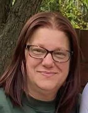 Smiling woman with glasses and brown hair outdoors near a tree.