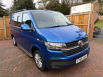 Blue Volkswagen van parked on a brick driveway near a wooden fence.