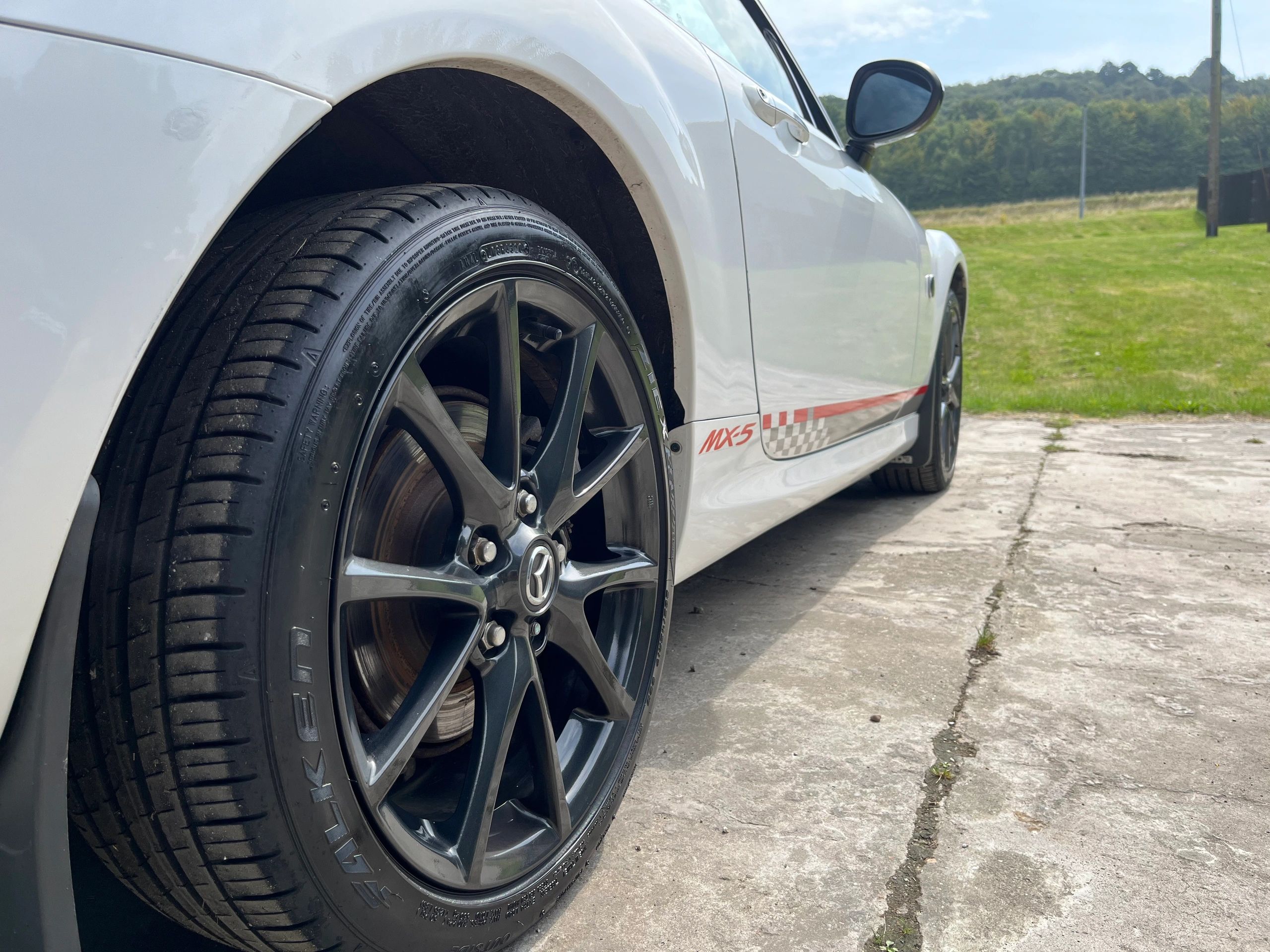 Close-up of a white Mazda MX-5 wheel and side panel on a sunny day.