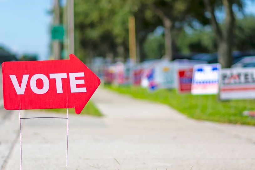 A red vote sign pointing along a sidewalk with campaign signs in the background.