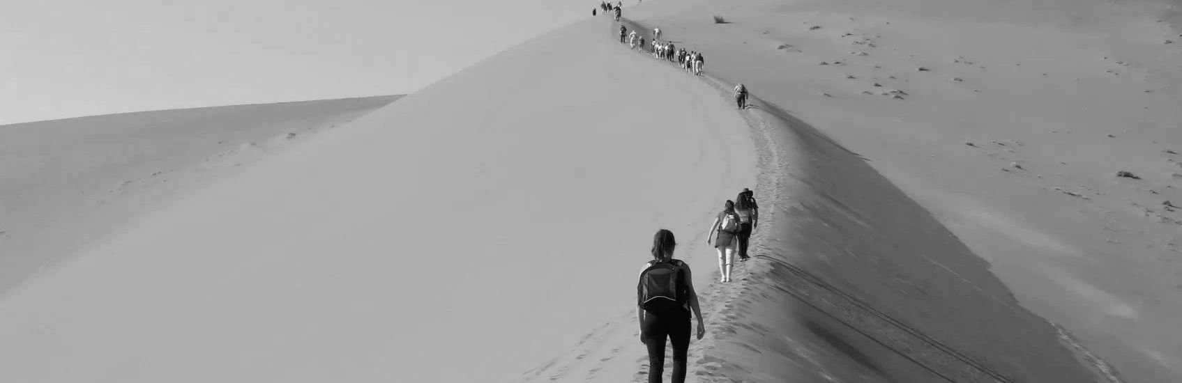 People hiking up a tall sand dune under a clear blue sky.