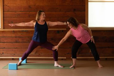 Two people practicing Warrior II pose with a block in a wood-lined studio.