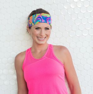 Smiling woman in bright pink athletic wear and colorful headband stands against a white textured wall.