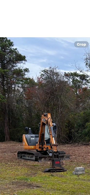 Yellow excavator on cleared land near a wooded area under a cloudy sky.