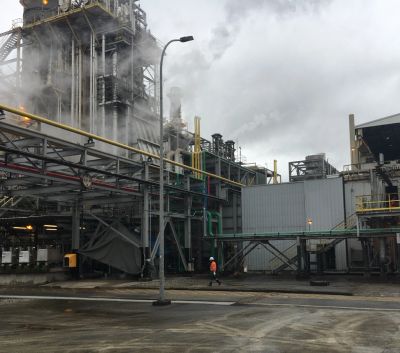 Industrial plant with conveyor belts and smoke, worker in orange vest.