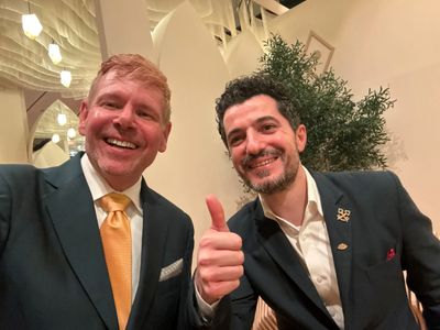 Two men in suits smiling, one giving a thumbs up indoors.