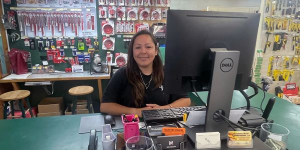 A woman smiling behind a store counter with tools and office supplies.