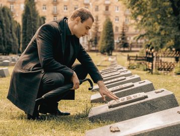 Man in black coat mourning at a cemetery gravestone.