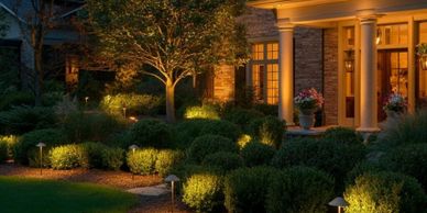 Warm outdoor lighting highlights a beautifully landscaped garden and house entrance at dusk.