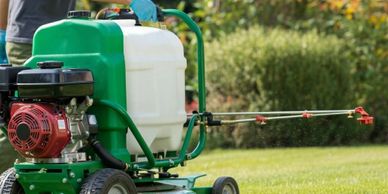 Person using a motorized sprayer on a green lawn with trees in the background.