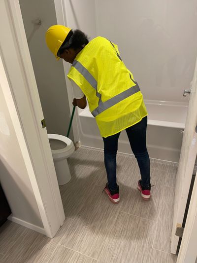 Person in a yellow safety vest cleaning a bathroom floor near a toilet.