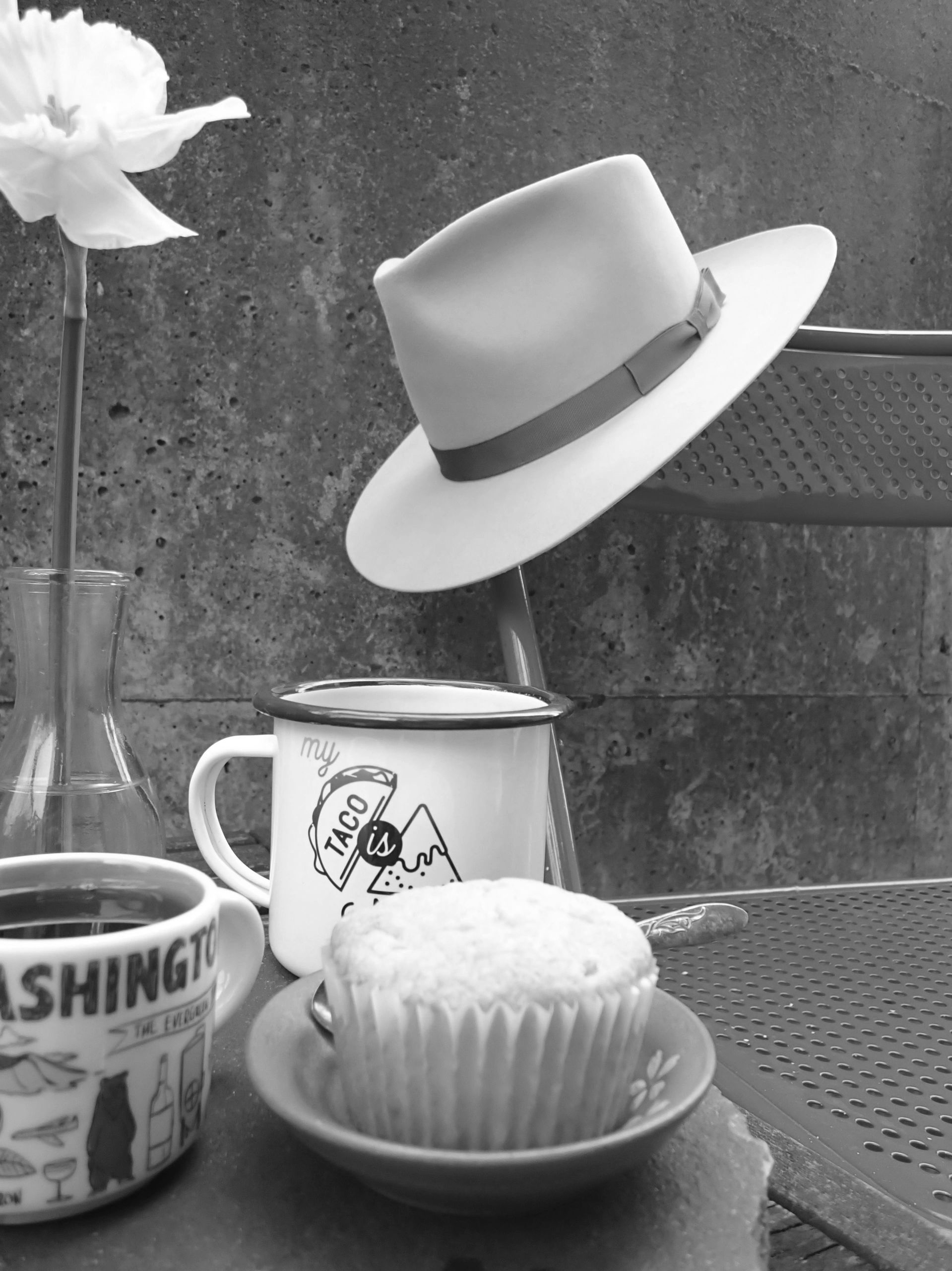 Coffee, a pastry, and a fresh-picked daffodil sit atop a table next to a fedora hanging on chair. 