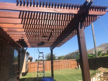 Wooden pergola casting patterned shadows over a backyard with a ladder and blue bucket.