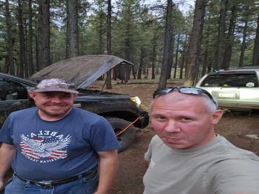 Two men smiling near a truck with its hood open in a forest.