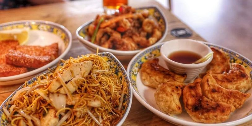 Plates of assorted Asian dishes including noodles, dumplings, and fried snacks on a wooden table.